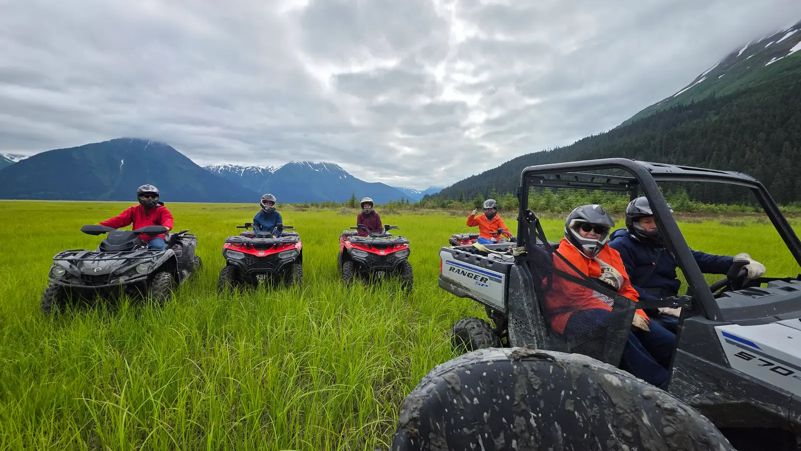 Five ATVs and a Ranger side-by-side lined up in tall green grass above Turnagain Arm, snow-streaked Chugach peaks behind