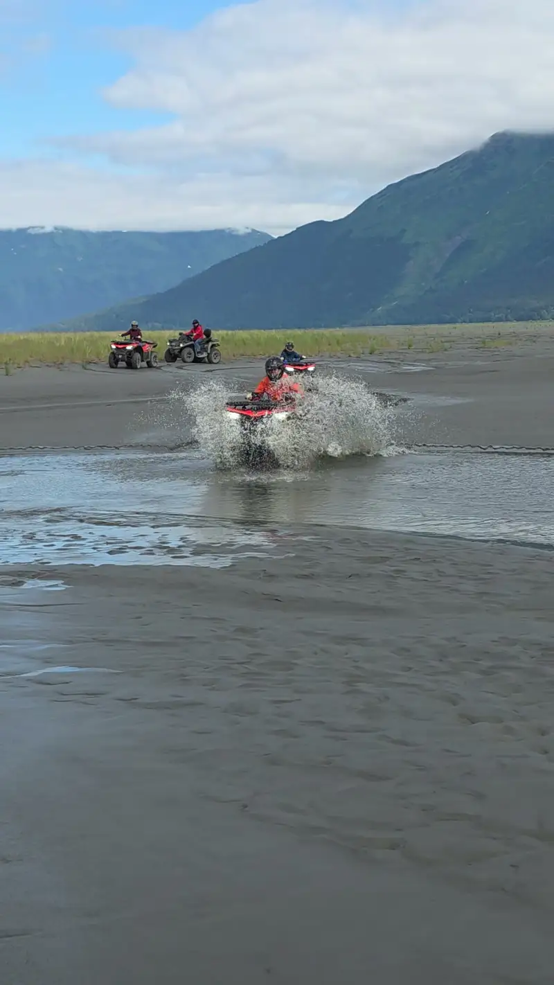 Red ATV driving straight at the camera through standing water on the tide flats, spray arcing to both sides