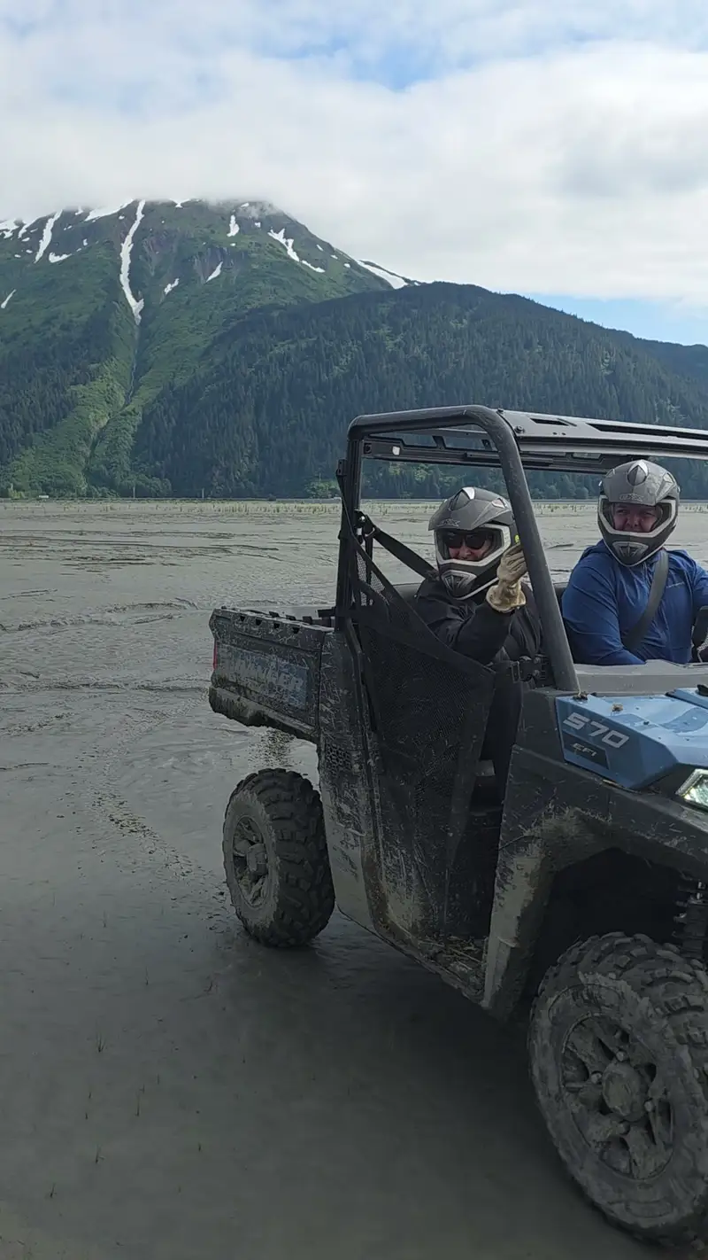 Two helmeted riders in a mud-caked Polaris Ranger on the Turnagain Arm flats, one giving a thumbs up