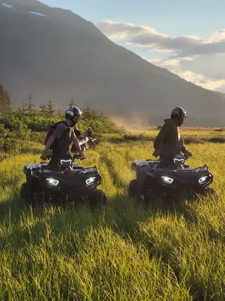 Two ATVs and their riders pausing in a grassy meadow above Turnagain Arm at golden hour