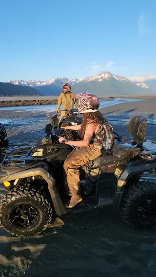 Two muddy riders and their ATVs after the AKMUDITUP tideland ride, inlet and mountains behind